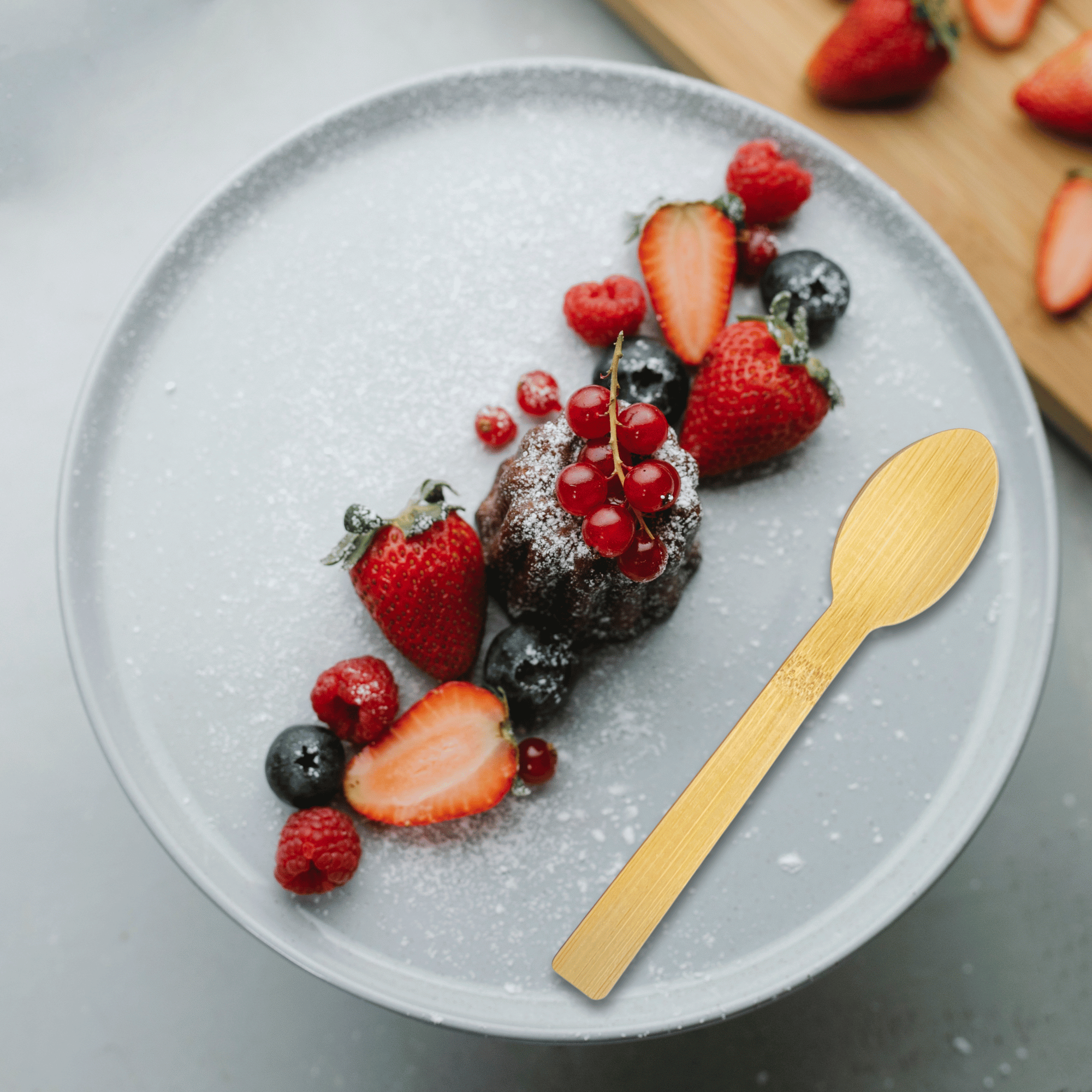 A bamboo spoon from Holy City Straw Co. lies next to a chocolate dessert sprinkled with powdered sugar, adorned with a sprig of red currants. The plate is artistically arranged with fresh strawberries, blueberries, raspberries, and slices of strawberries, on a light grey ceramic plate with a soft-focus wooden surface in the background.