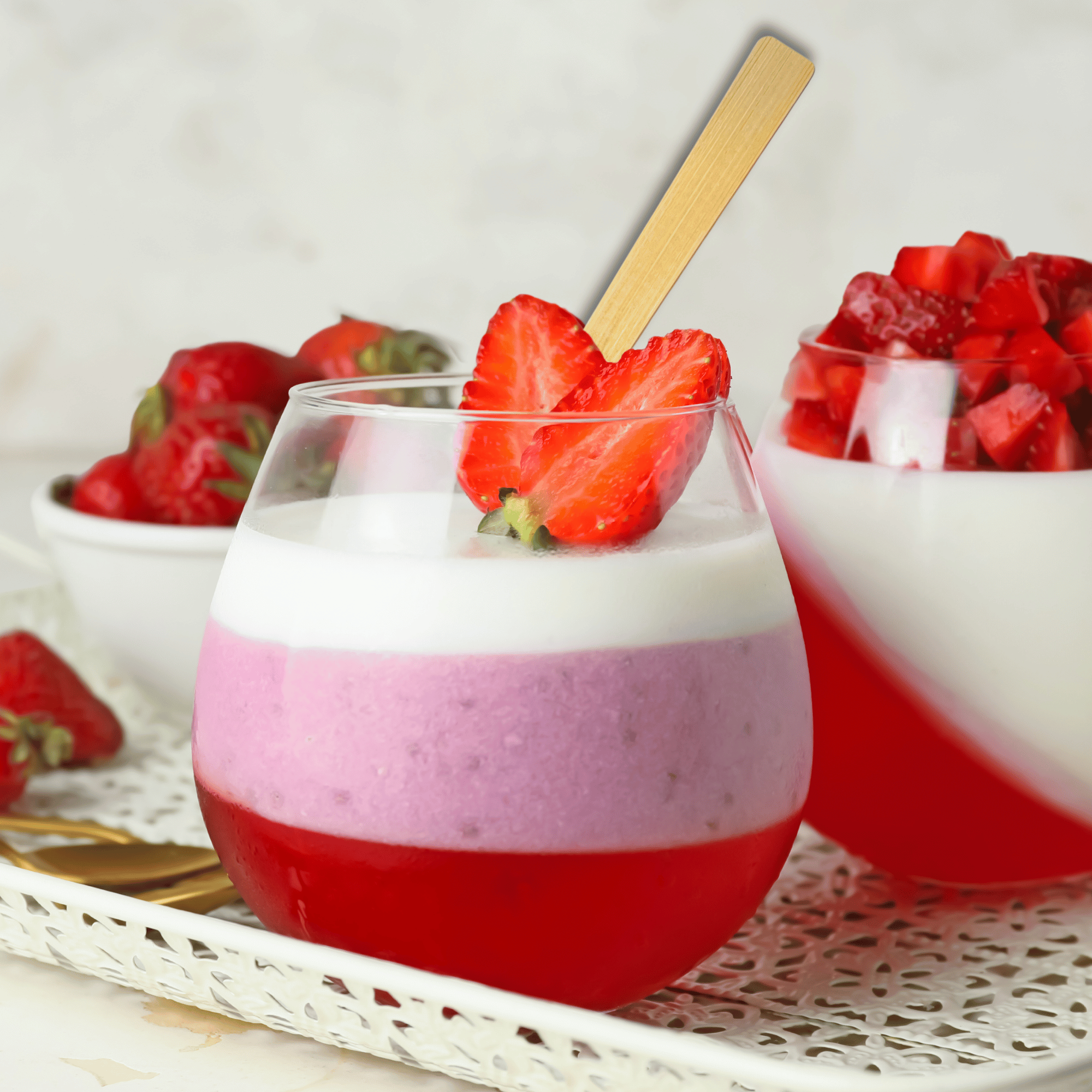 An elegant dessert presented in a clear glass showcases layered strawberry jelly and creamy purple mousse topped with a fluffy white cream. A sliced strawberry garnishes the rim and a bamboo spoon from Holy City Straw Co. is inserted, ready for indulgence.