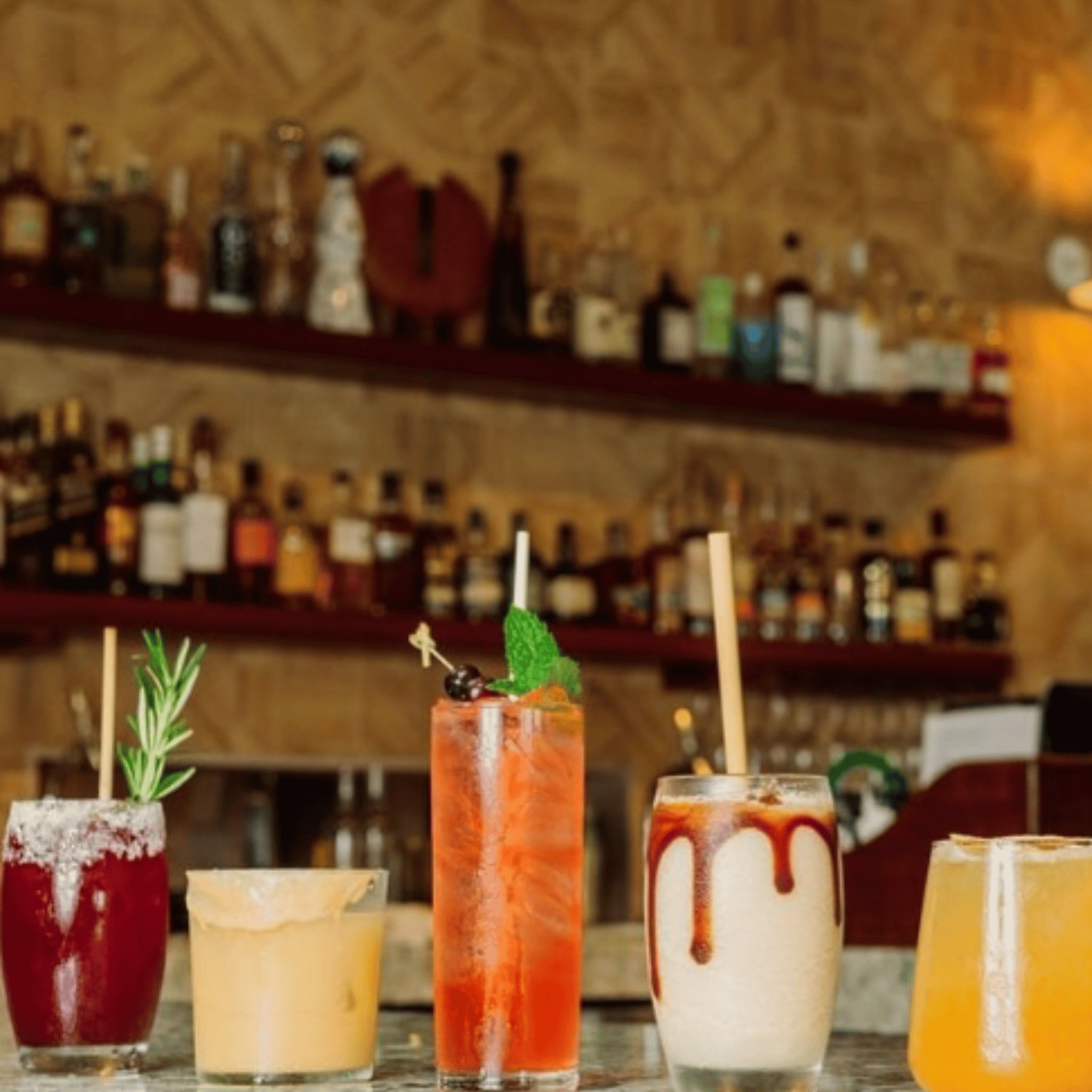 A variety of five colorful cocktails on a bar counter including a margarita with a salt rim, a tall glass of red sangria with a fruit garnish, a long drink with a sprig of rosemary, a creamy cocktail topped with a chocolate swirl, and a bright yellow drink in a short glass. Each cocktail features a different type of garnish, emphasizing their unique flavors and presentations. In the background, an extensive collection of liquor bottles is displayed on wooden shelves, enhancing the ambiance.