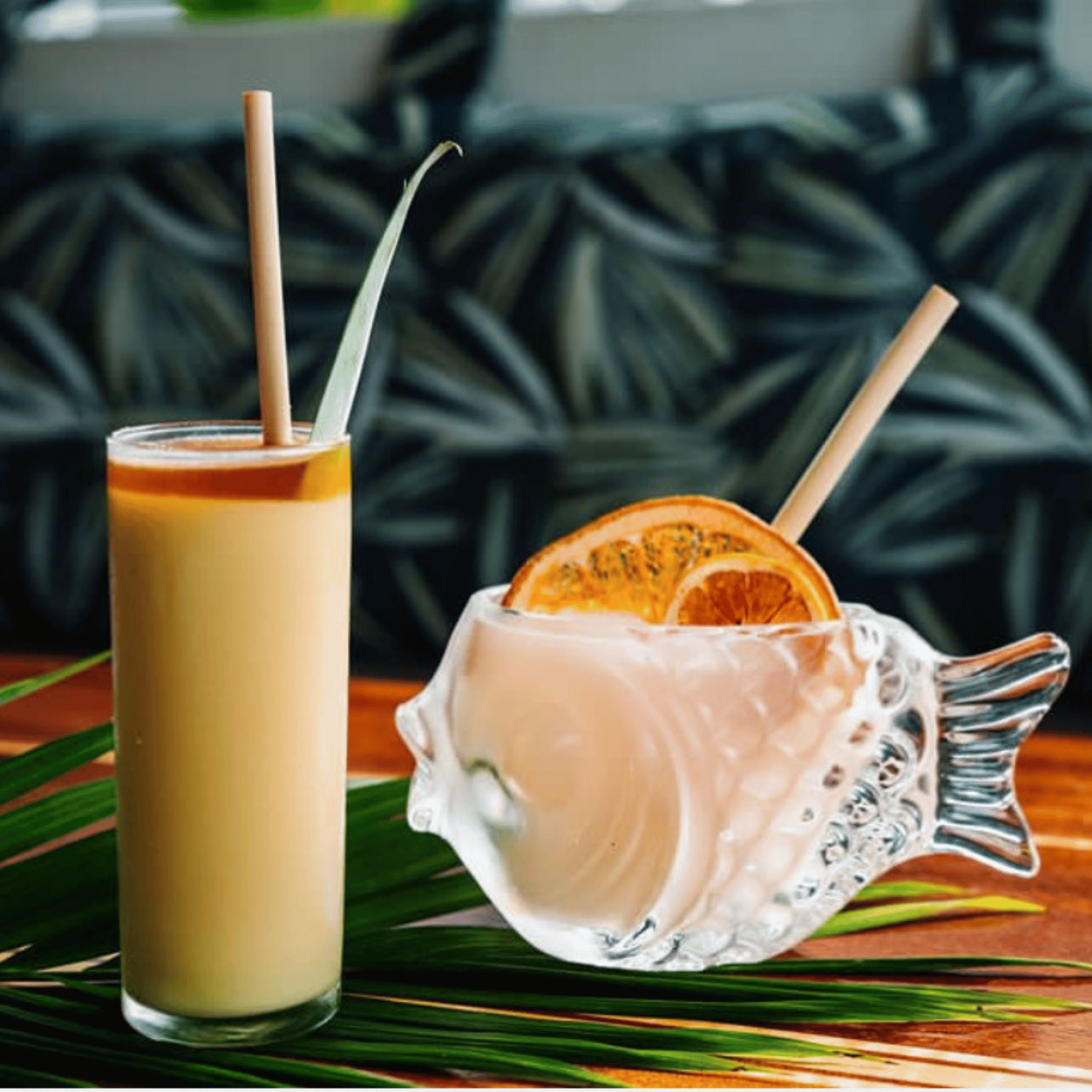 A tall glass of frozen rum drink with a bamboo straw beside a unique fish-shaped glass filled with a creamy pink cocktail, garnished with a dried orange slice, both on a palm leaf against a tropical patterned backdrop