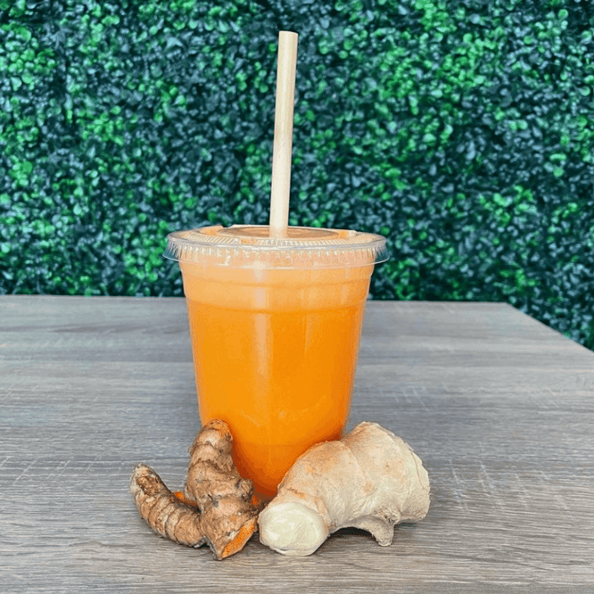 A bright orange juice blend in a clear plastic cup with a bamboo straw, shown on a wooden table alongside fresh ginger and turmeric roots against a lush green wall.