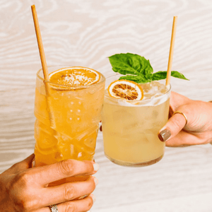 Two people are toasting with cocktails featuring skinny reed straws made from reed stems. The drinks are garnished with dried citrus slices and fresh herbs, emphasizing a sustainable and refreshing aesthetic against a light wooden backdrop.