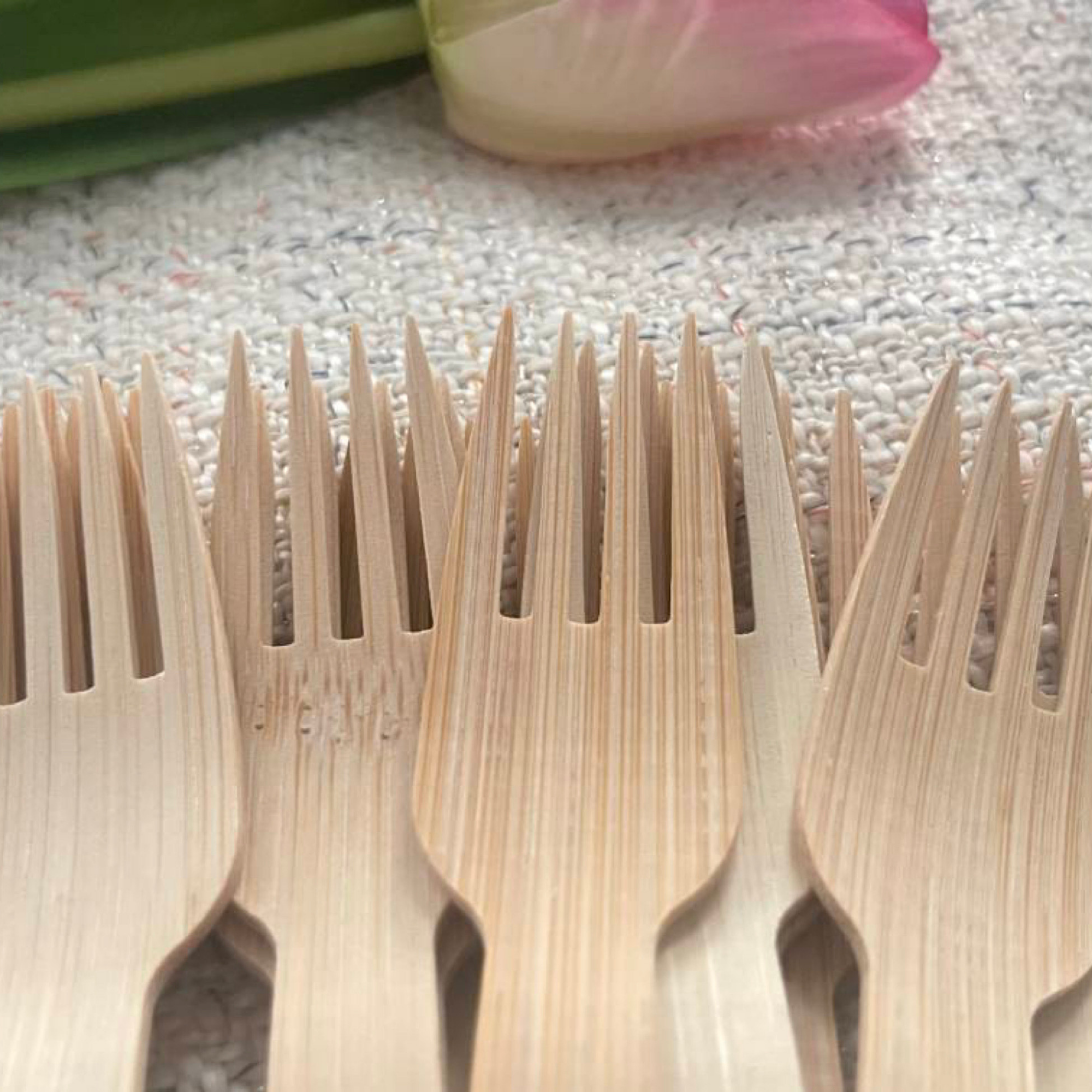 Close-up of Holy City Straw Co. bamboo forks, showcasing the finely detailed tines and natural wood texture. The forks are grouped together on a textured fabric, with a soft-focus view of a pink tulip and fresh produce in the background, highlighting the brand's commitment to nature and sustainability.