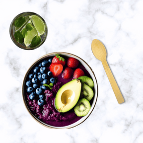 An aesthetically pleasing top-view of a vibrant smoothie bowl from Holy City Straw Co., decorated with an assortment of fresh fruits including sliced strawberries, kiwi, avocado, and a generous handful of blueberries. Accompanied by a refreshing glass of lime-infused water with mint leaves, and a sustainable wooden spoon positioned to the right, set on a chic marble background.