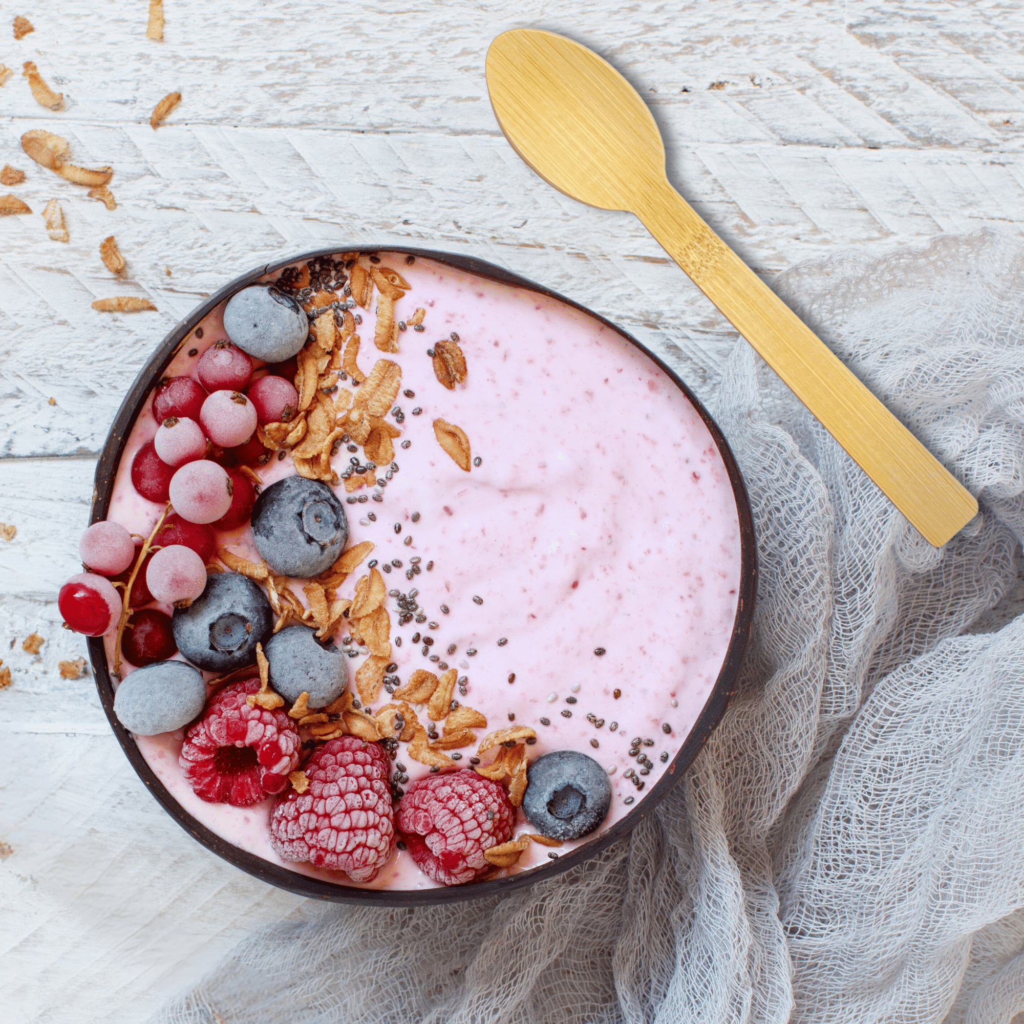 An appealing top-view image showcasing Holy City Straw Co.'s latest sustainable cutlery, a wooden spoon resting on the side of a bowl filled with creamy pink smoothie. The bowl is beautifully topped with a variety of frozen fruits including red grapes, blueberries, and raspberries, sprinkled with granola and chia seeds, all arranged on a rustic white wooden surface with a delicate gauze fabric underneath.