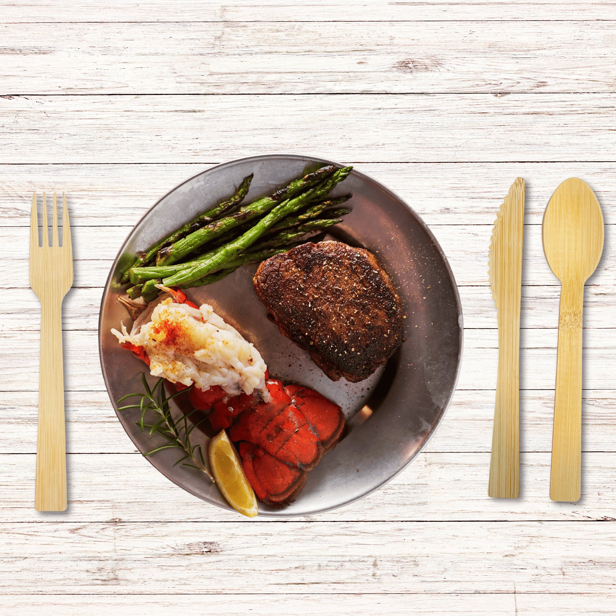 An elegant dinner setting featuring Holy City Straw Co. brand eco-friendly wooden cutlery, with a fork to the left and knife and spoon to the right. The meal includes a well-seasoned steak, succulent lobster tail, a scoop of creamy crab meat, and a side of grilled asparagus, all artistically arranged on a round plate with a rustic wooden table background