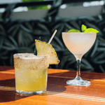 A pineapple cocktail with a salted rim sits beside a martini glass filled with a clear drink garnished with mint. Both drinks are placed on a wooden surface, and the pineapple cocktail includes a skinny reed straw. The background features palm-patterned upholstery, providing a vibrant tropical feel.