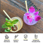 Two colorful cocktails sit on a wooden surface. One is garnished with mint leaves and a dried citrus slice, while the other has a purple orchid flower. Both glasses contain cocktail skinny reed straws made from reed stems. Icons at the bottom emphasize that these straws are made from reed stems, similar to bamboo, durable, and designed for everyday drinks.