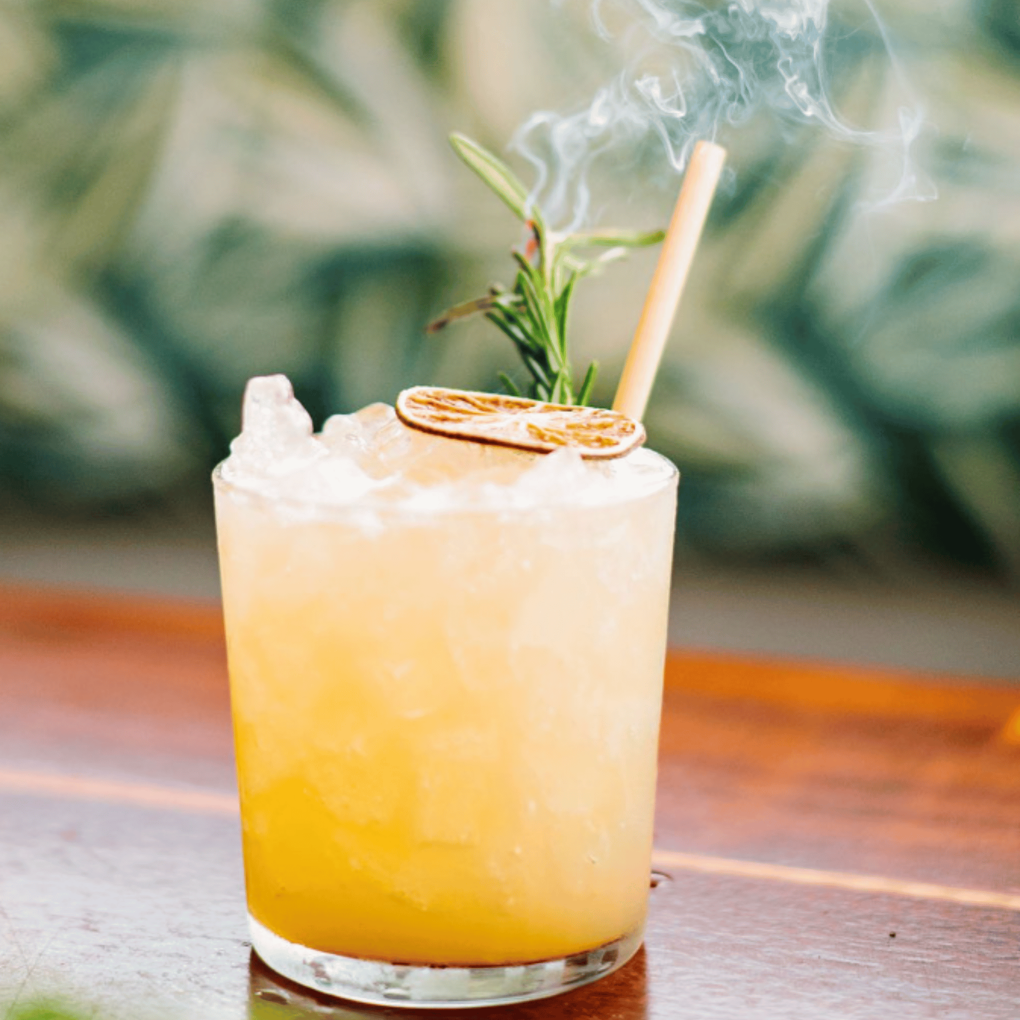  An iced cocktail garnished with a sprig of rosemary and a dried citrus slice sits on a wooden table. The glass has a skinny reed straw, and smoke from the herb adds a touch of flair, highlighting a lush green background.