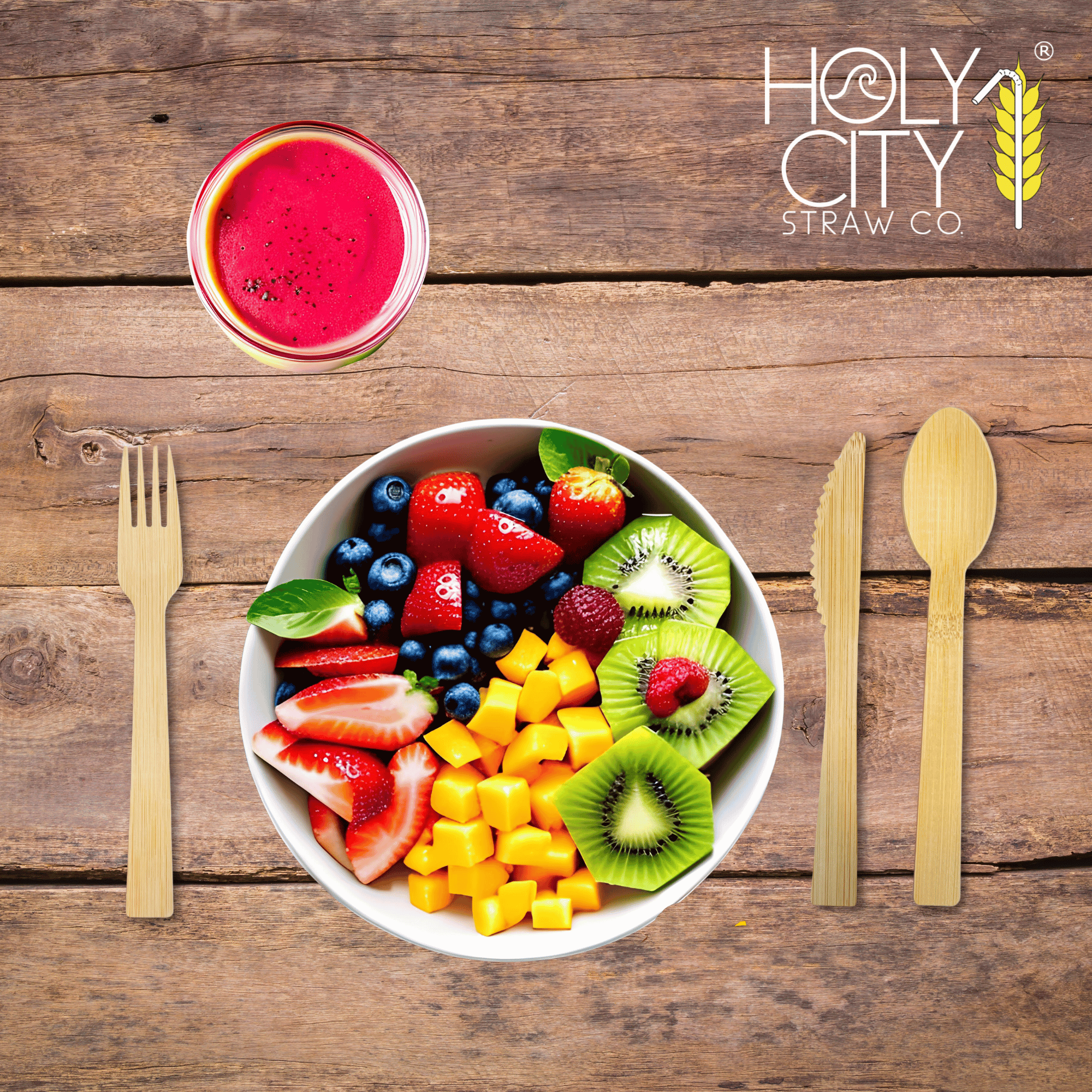 Top view of a wooden table setting featuring Holy City Straw Co. brand bamboo cutlery, with a bamboo fork on the left and a knife on the right, framing a colorful bowl of fresh fruit salad containing strawberries, blueberries, kiwi, and mango cubes. Above the bowl is a glass of red smoothie, and the Holy City Straw Co. logo with a wheat stalk icon is prominently displayed at the top.