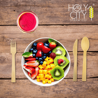 Top view of a wooden table setting featuring Holy City Straw Co. brand bamboo cutlery, with a bamboo fork on the left and a knife on the right, framing a colorful bowl of fresh fruit salad containing strawberries, blueberries, kiwi, and mango cubes. Above the bowl is a glass of red smoothie, and the Holy City Straw Co. logo with a wheat stalk icon is prominently displayed at the top.