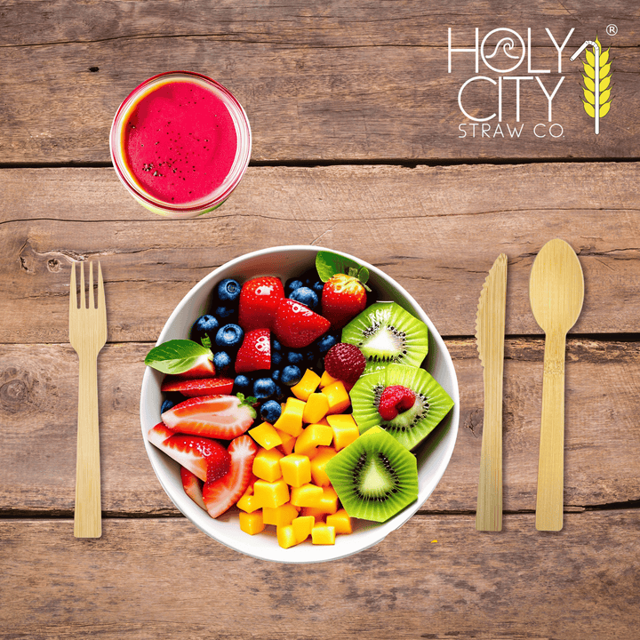 Top view of a wooden table setting featuring Holy City Straw Co. brand bamboo cutlery, with a bamboo fork on the left and a knife on the right, framing a colorful bowl of fresh fruit salad containing strawberries, blueberries, kiwi, and mango cubes. Above the bowl is a glass of red smoothie, and the Holy City Straw Co. logo with a wheat stalk icon is prominently displayed at the top