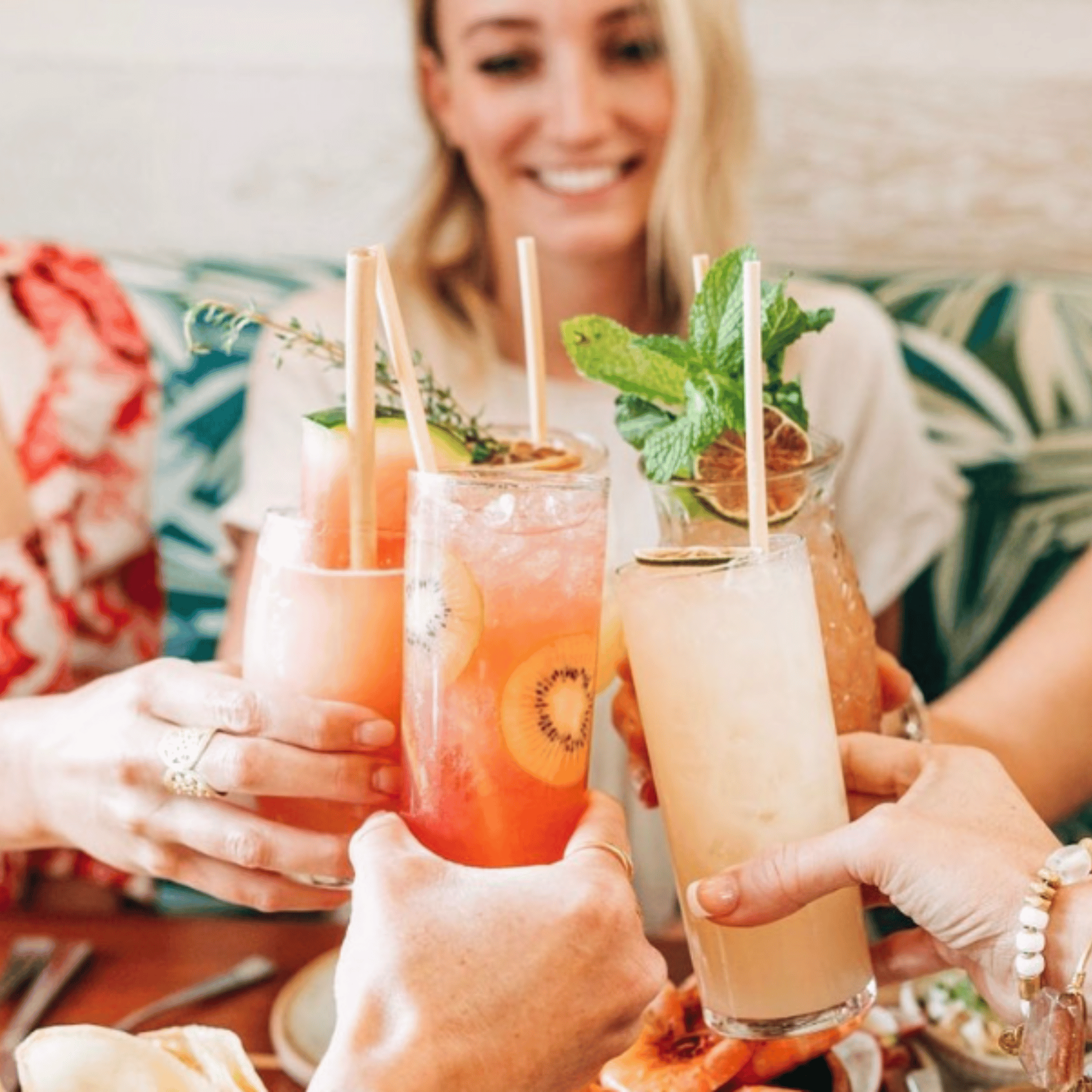 Four friends raise their cocktails in a cheerful toast. Each drink is garnished with fresh fruit or herbs, featuring tall skinny reed straws for an eco-friendly touch. The background includes a person smiling and tropical upholstery, contributing to the festive and relaxed atmosphere.