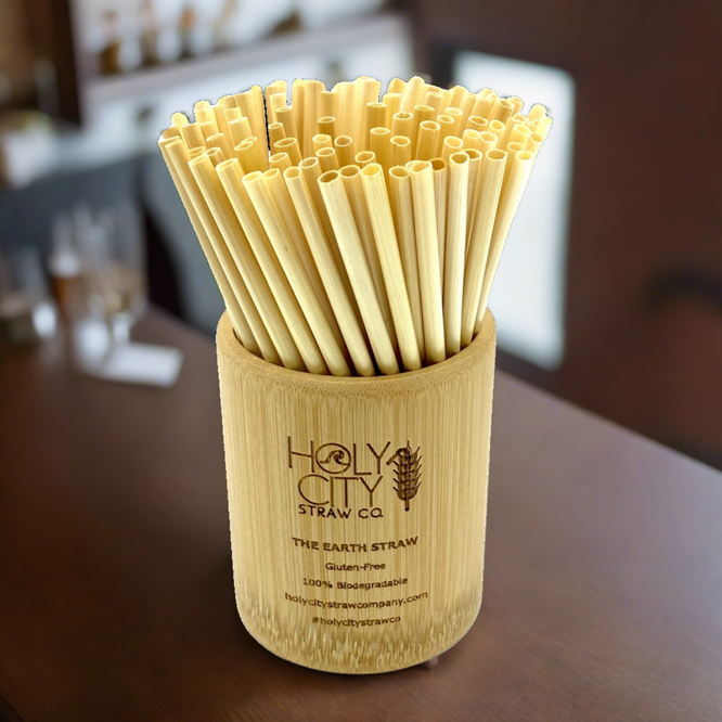 A cylindrical bamboo holder branded with ‘HolyCity Straw Co. – The Earth Straw,’ filled with a large bundle of natural bamboo straws. The holder’s etched text mentions gluten-free, biodegradable features, and it rests on a blurred tabletop background.