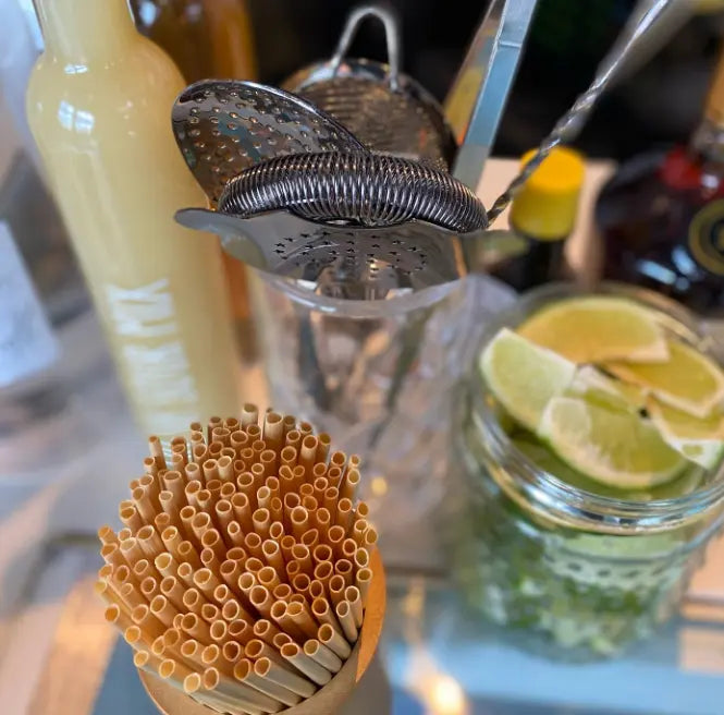 Eco-friendly straws from Holy City Straw Co. in a wooden holder, placed next to cocktail-making tools, lime wedges in a jar, and a bottle of mixer on a bar counter.