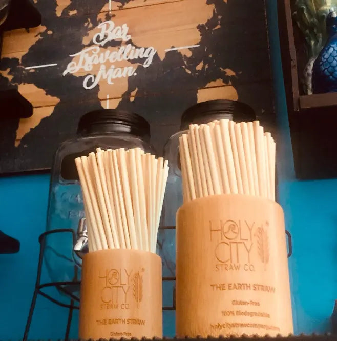 Wooden holders filled with eco-friendly straws from Holy City Straw Co., displayed on a counter with glass jars and a 'Big Travelling Man' sign in the background.