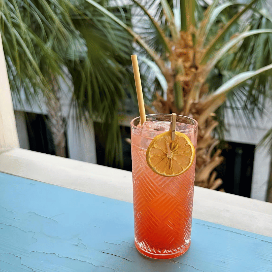 A refreshing pink cocktail in a tall, textured glass with a lemon slice garnish and bamboo straws, sitting on a weathered blue table. Palm trees and a building are visible in the background, suggesting a tropical, outdoor setting on March 3, 2025

