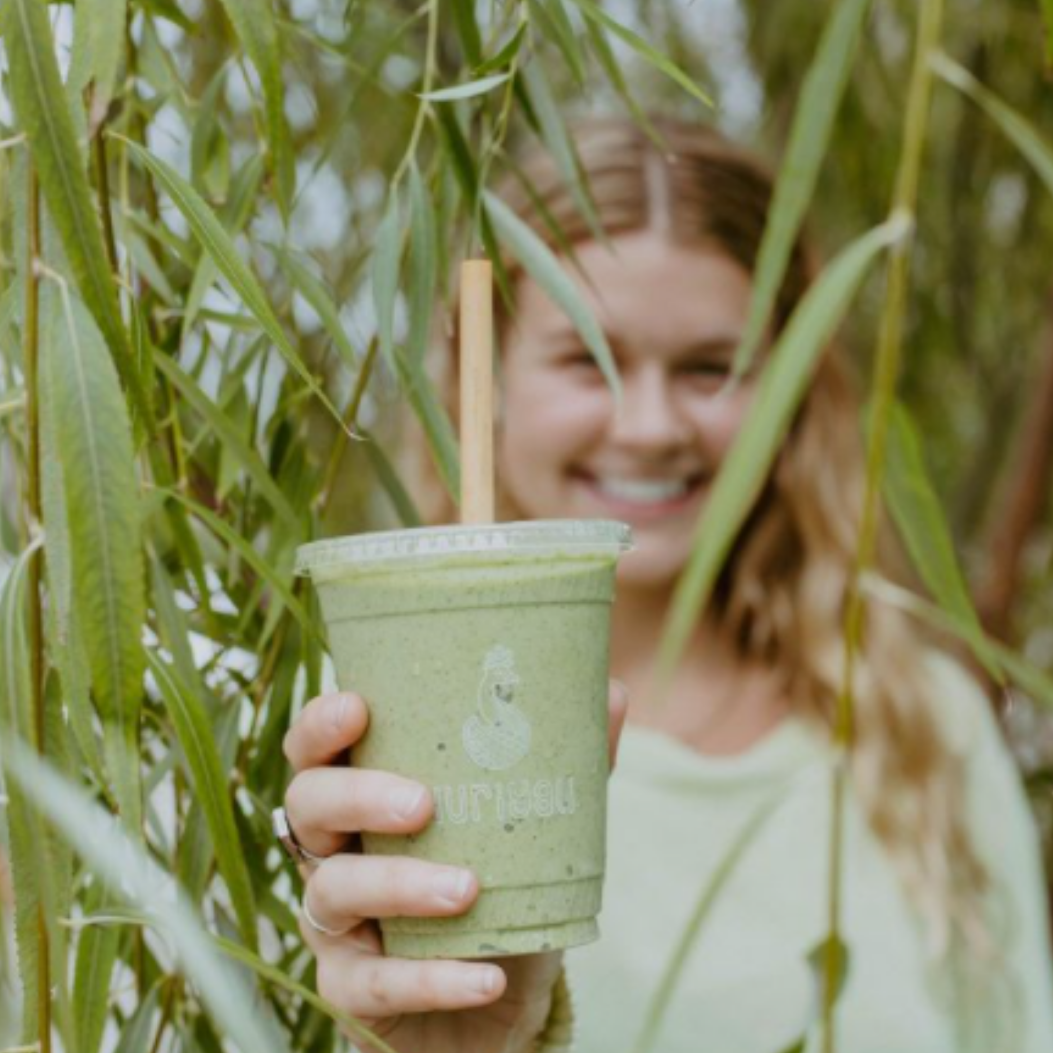 Girl holding a green smoothie through palm trees highlighting the reed straw