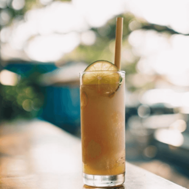 A tall, iced beverage in a clear glass garnished with lime slices and featuring a bamboo straw, set against a softly blurred outdoor backdrop.