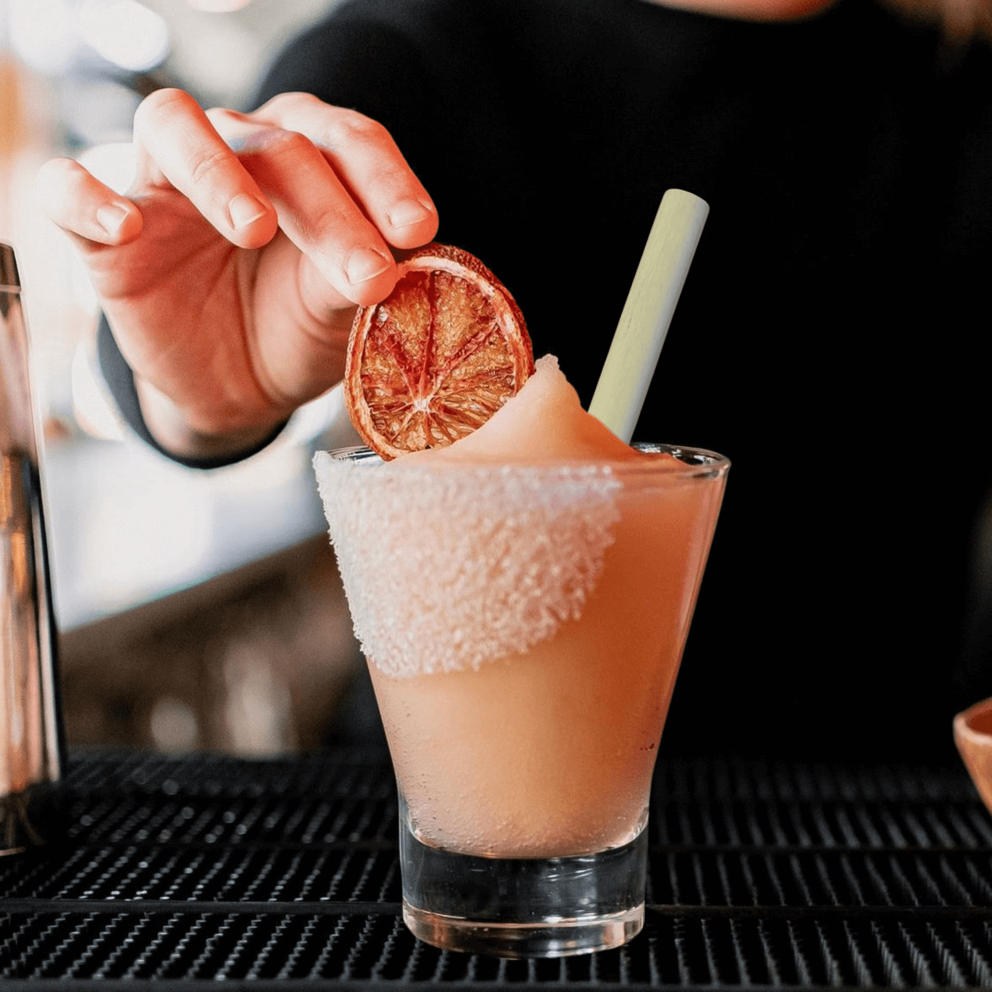 A hand is placing a dried grapefruit slice as a garnish on a frozen paloma served in a glass with a salted rim. The glass sits on a dark bar surface and contains a reed straw, which resembles bamboo for an eco-friendly touch. The background is blurred, emphasizing the drink's colorful and stylish presentation, highlighting the sustainability aspect of the straw.