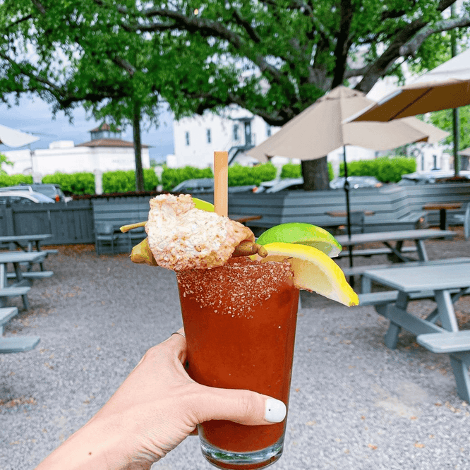 A chili-rimmed Bloody Mary held in someone’s hand, topped with lime and lemon wedges, a crunchy garnish, and a bamboo straw in an outdoor patio setting.