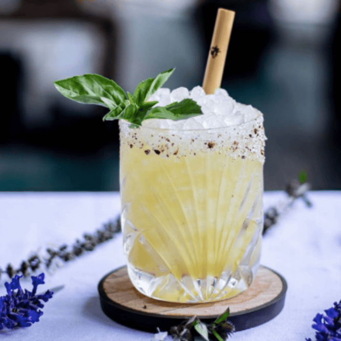 A pale yellow cocktail in an ornate crystal glass, filled with crushed ice and topped with fresh basil leaves. A bamboo straw stands in the drink, and the rim has a light seasoning around the edge.