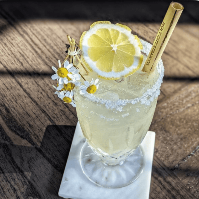 A tall lemon-yellow cocktail in an ornate glass rimmed with sugar, garnished with a fresh lemon slice and small daisies. A bamboo straw printed with ‘Holy City Straw Co.’ stands in the icy drink on a wooden table