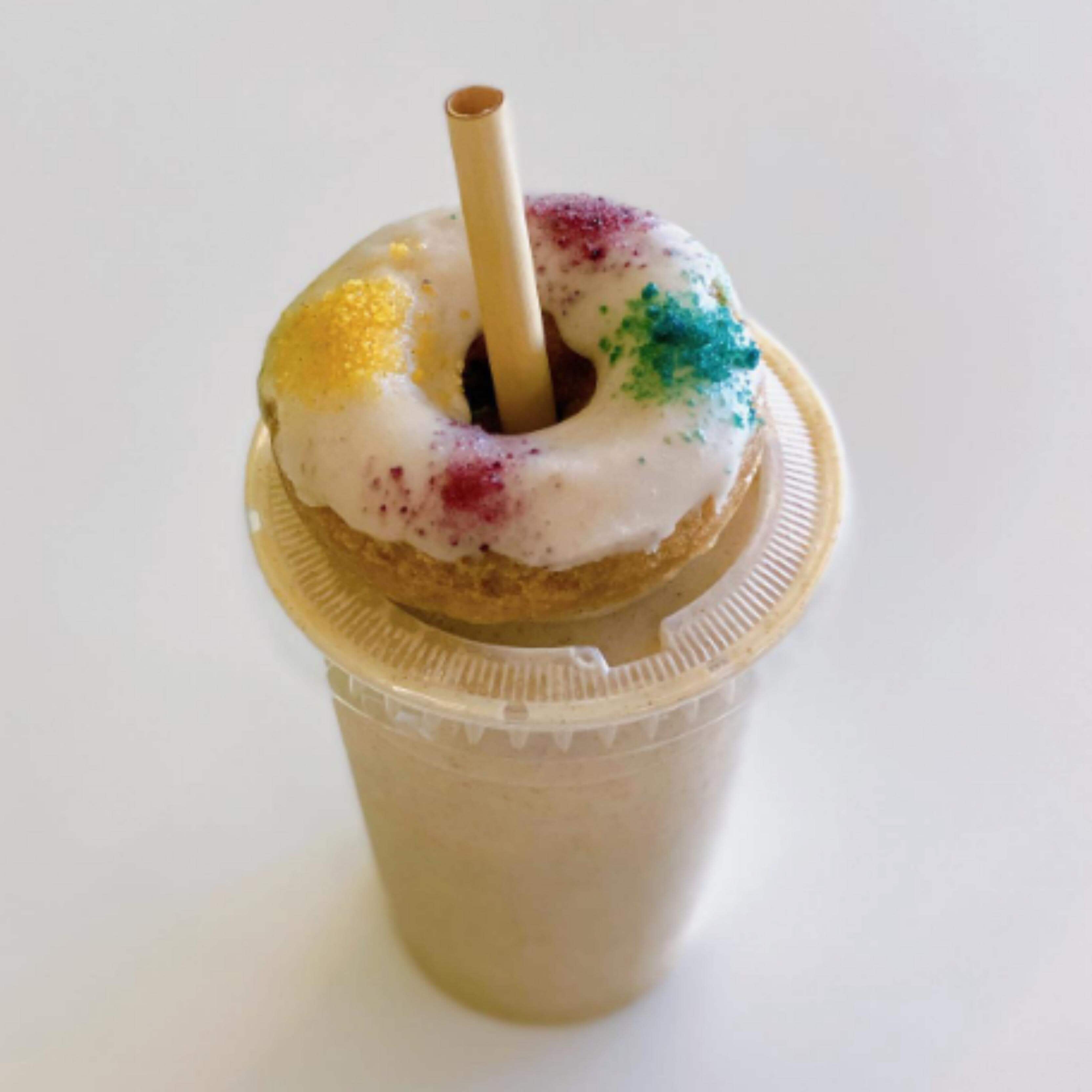 A milkshake in a clear cup is topped with a colorful donut, which has a jumbo reed straw inserted through its center. The straw, made from biodegradable reed stems and resembling bamboo, provides a sustainable touch. The donut is decorated with a white icing glaze and multicolored sugar crystals, creating a playful, whimsical garnish. The background is plain white, emphasizing the focus on the drink's unique presentation.
